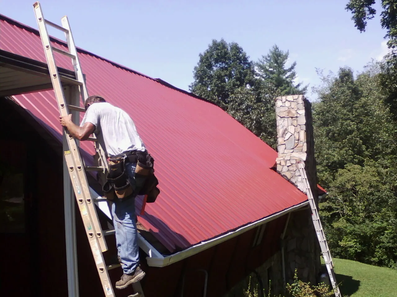 Roofer on a ladder beside a red metal roof and stone chimney — Soffit & Fascia Repair by Roofing Brothers in Pitman